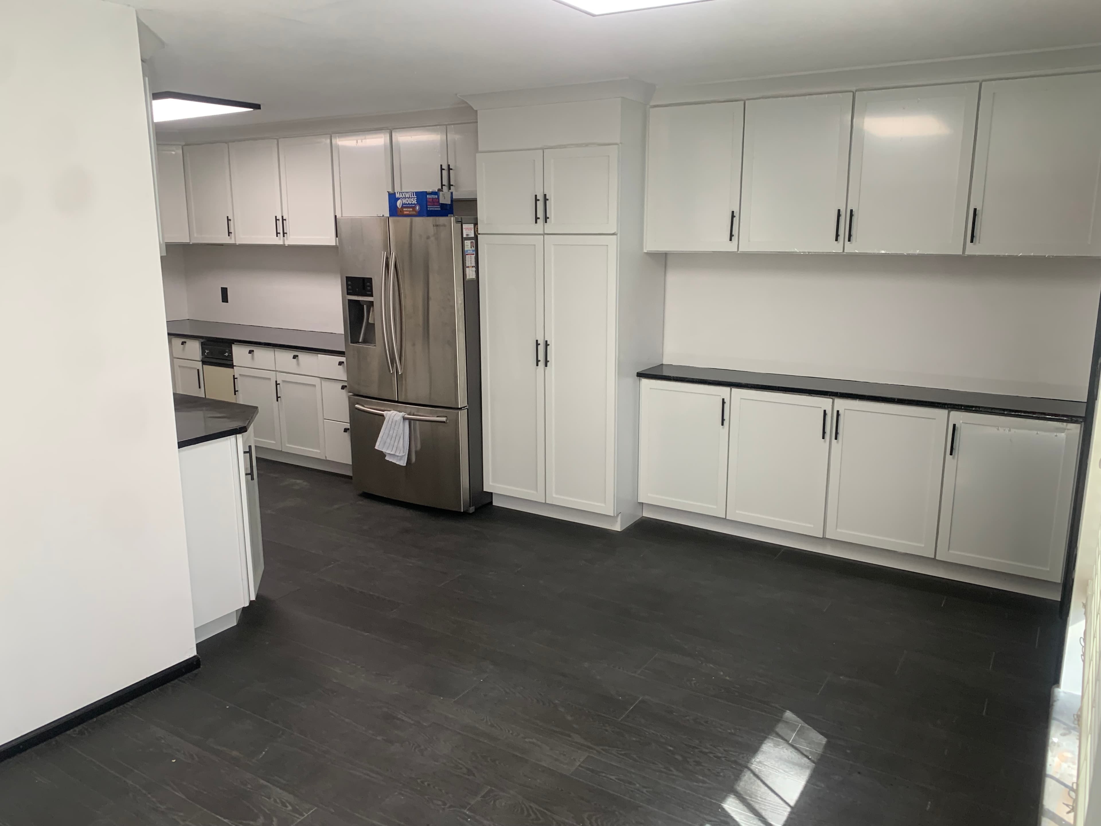 Modern white kitchen with black countertops, dark wood-look flooring, and a stainless steel refrigerator.