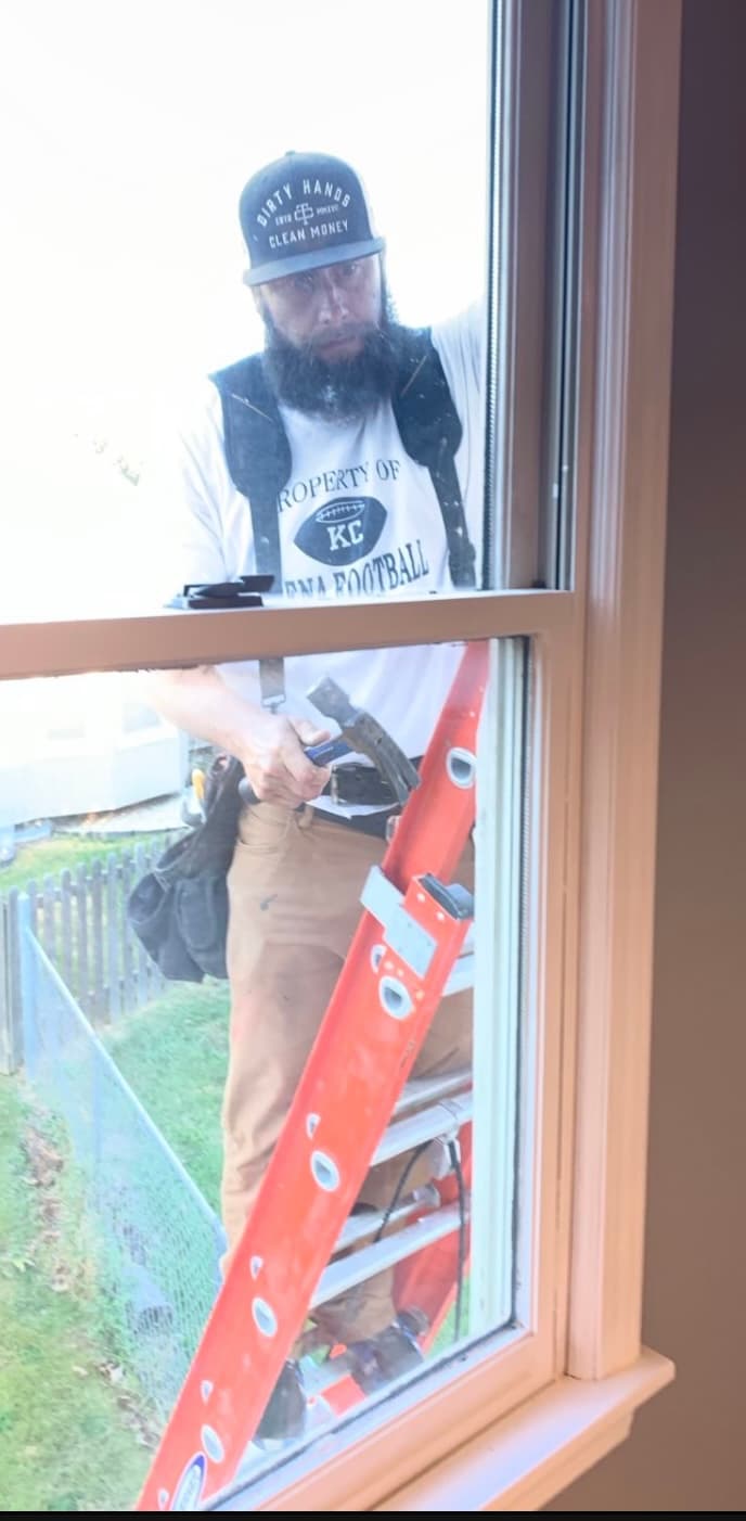 Bearded man on an orange ladder holds a hammer while working on a white window.